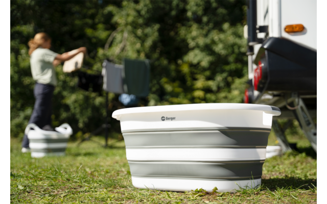 Faltbarer weiß-grauer Waschbehälter im Vordergrund, im Hintergrund eine Person, die Wäsche an einem Wäscheständer neben einem Wohnwagen aufhängt