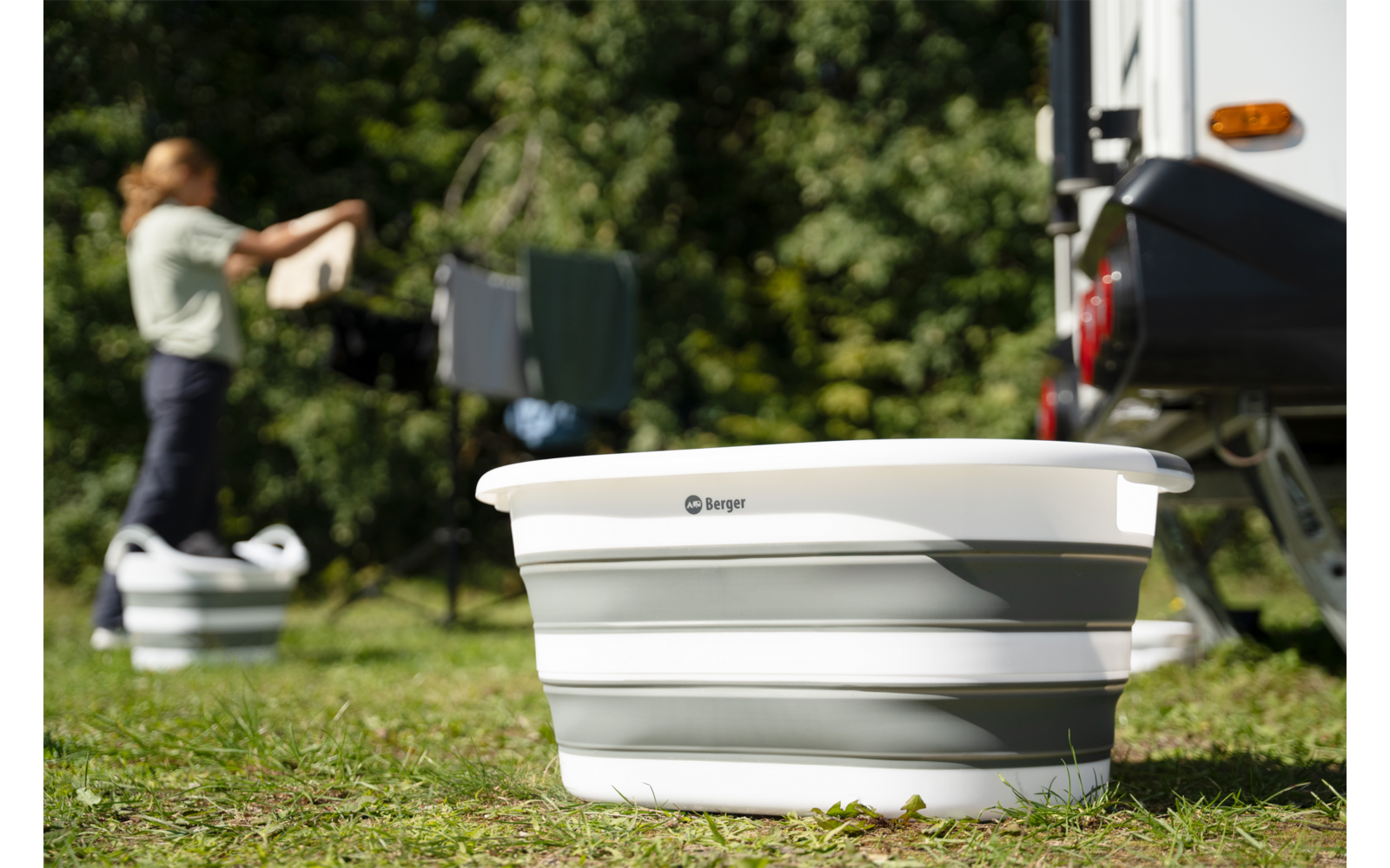 Faltbarer weiß-grauer Waschbehälter im Vordergrund, im Hintergrund eine Person, die Wäsche an einem Wäscheständer neben einem Wohnwagen aufhängt