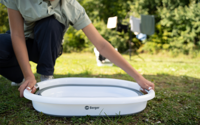 Eine Person kniet auf Gras und stellt eine weiße, zusammenklappbare Waschschüssel mit Aufdruck 'Berger' ab; im Hintergrund Wäsche an einer Leine und Bäume