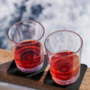 Zwei Gläser mit roter Flüssigkeit auf einem Holzbrett mit schwarzem Untersetzer, unscharfer Hintergrund mit Wasser