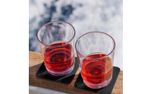 Two glasses with red liquid on a wooden board with black coasters, blurred background with water