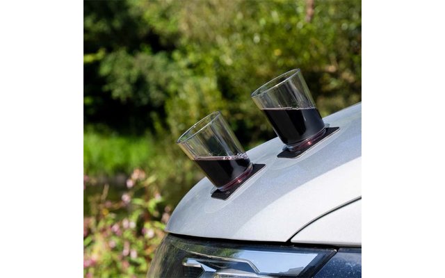 Zwei transparente Gläser mit dunkler Flüssigkeit auf schwarzen Untersetzern auf der Motorhaube eines silbernen Autos, unscharfer grüner Hintergrund