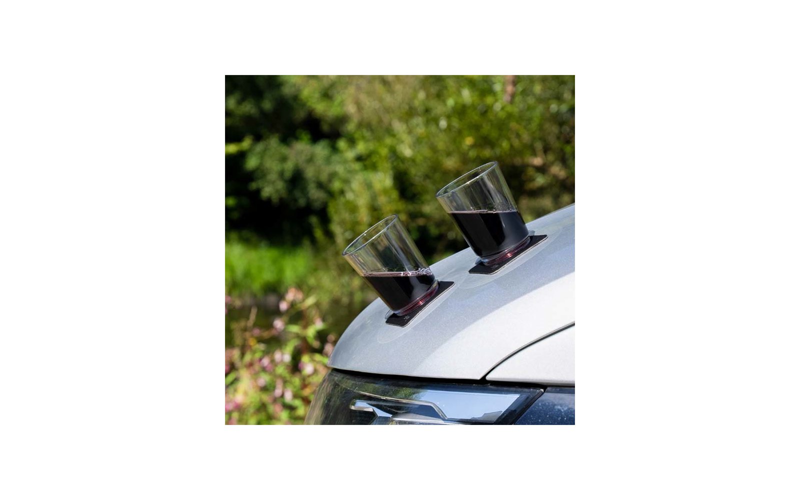 Zwei transparente Gläser mit dunkler Flüssigkeit auf schwarzen Untersetzern auf der Motorhaube eines silbernen Autos, unscharfer grüner Hintergrund