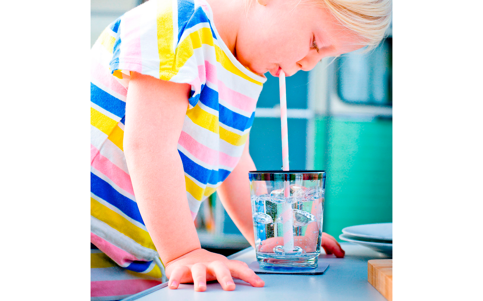 Kind in farbig gestreiftem T-Shirt trinkt Wasser aus einem Glas mit Strohhalm