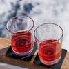 Zwei Glasbecher mit rotem Getränk auf einem Holzbrett vor unscharfem Hintergrund