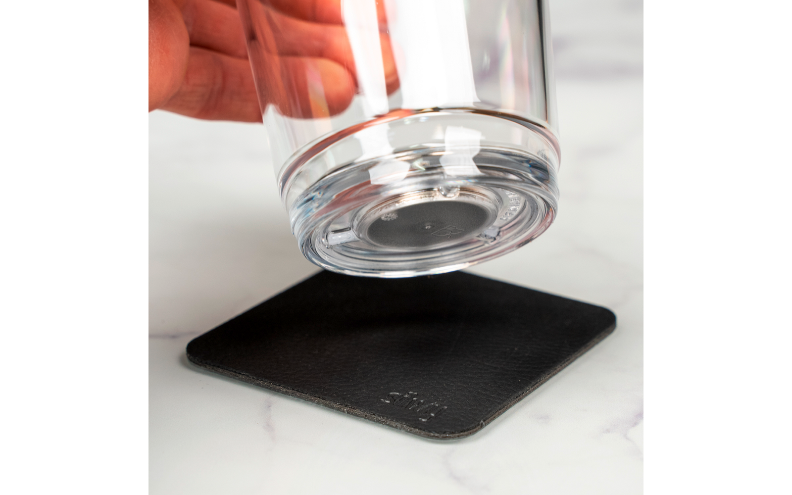 Hand hält ein leeres Glas über einen schwarzen Untersetzer auf weißer Oberfläche