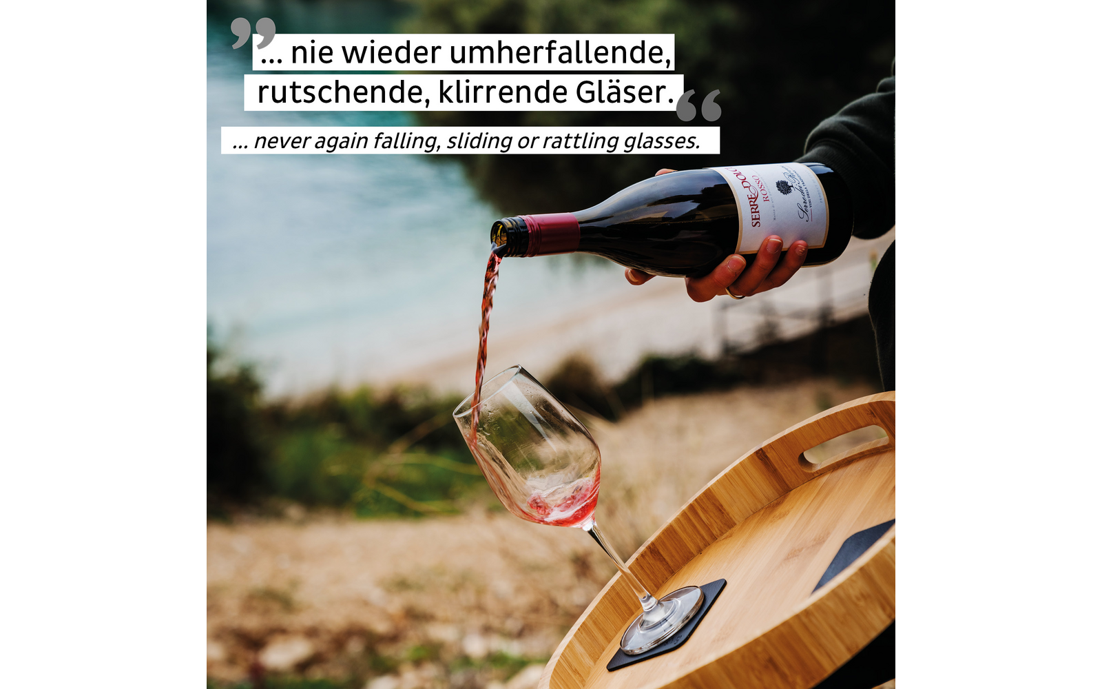 Person gießt roten Wein aus einer Flasche in ein Glas, das auf einem runden Holztablett steht, draußen am Wasser