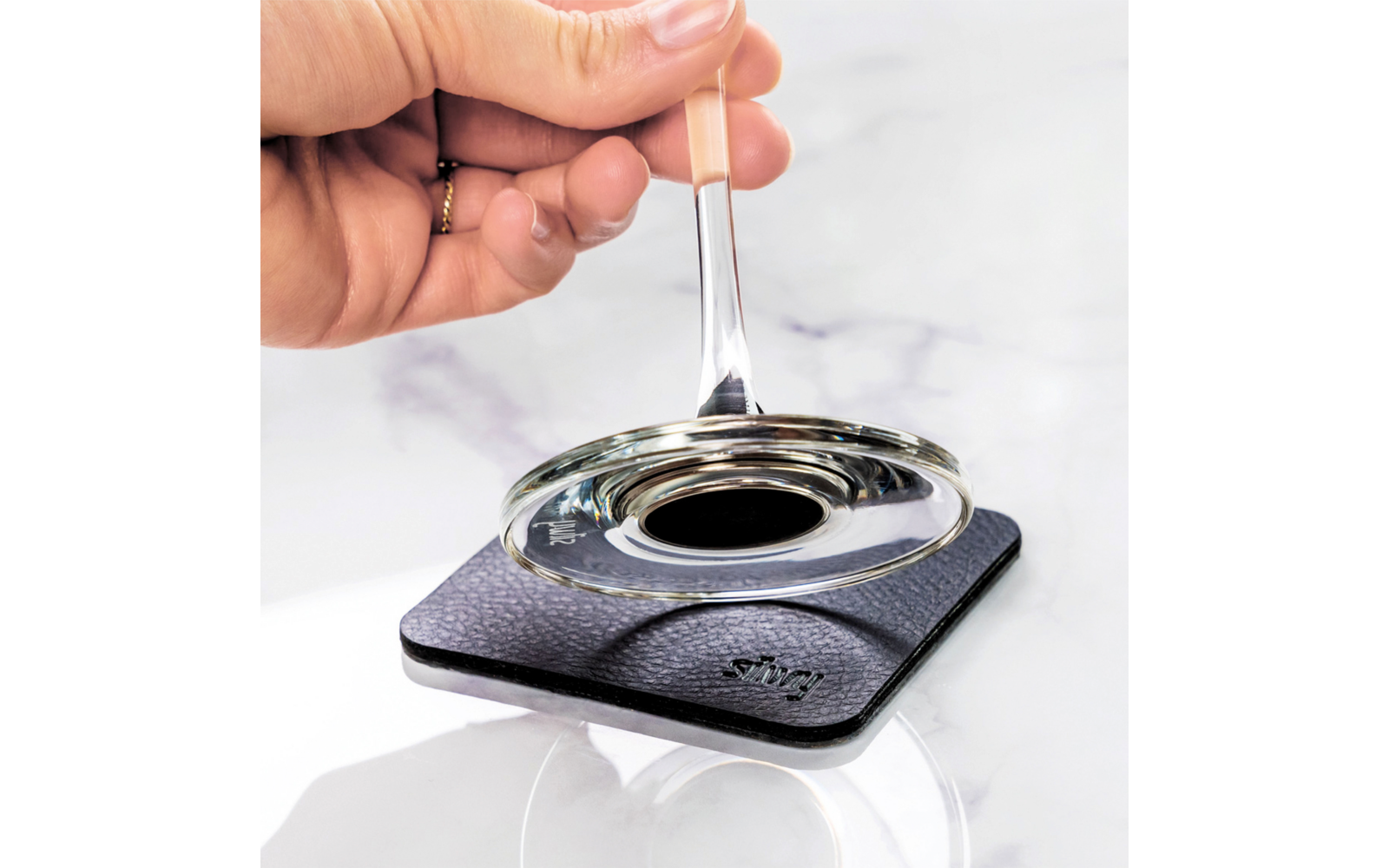 Hand with ring holding a glass rod over a round glass dish on a square black coaster on a marble surface
