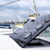 Two wine glasses with white wine on a tilted dark glossy solar panel of a boat hatch; background shows water, sailboat masts and cloudy sky