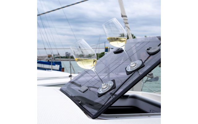 Two wine glasses with white wine on a tilted dark glossy solar panel of a boat hatch; background shows water, sailboat masts and cloudy sky