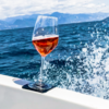 Stemmed glass with pink liquid on the edge of a boat, background sea, splashing water and mountains
