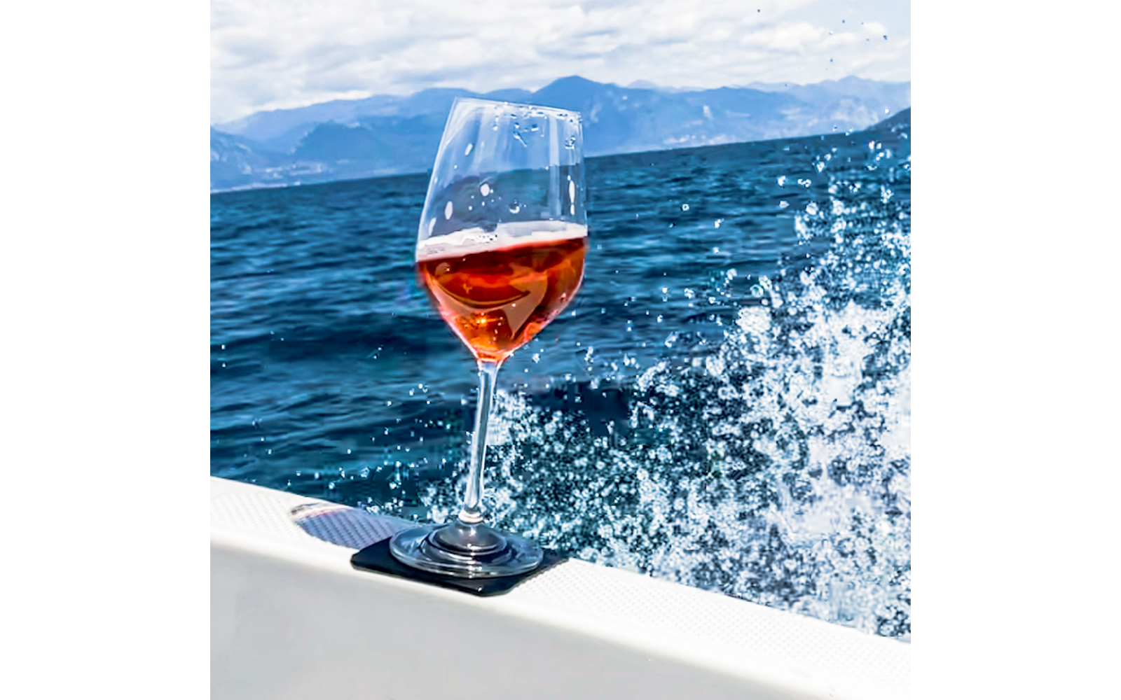 Stemmed glass with pink liquid on the edge of a boat, background sea, splashing water and mountains