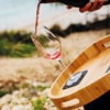 Hand pouring red liquid from a bottle into a tilted wine glass on a round wooden tray; blurred beach in the background