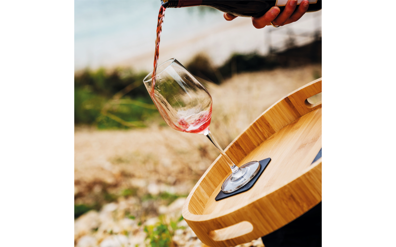 Hand pouring red liquid from a bottle into a tilted wine glass on a round wooden tray; blurred beach in the background