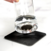 Ein Glas mit Wasser wird über einem schwarzen quadratischen Untersetzer gehalten