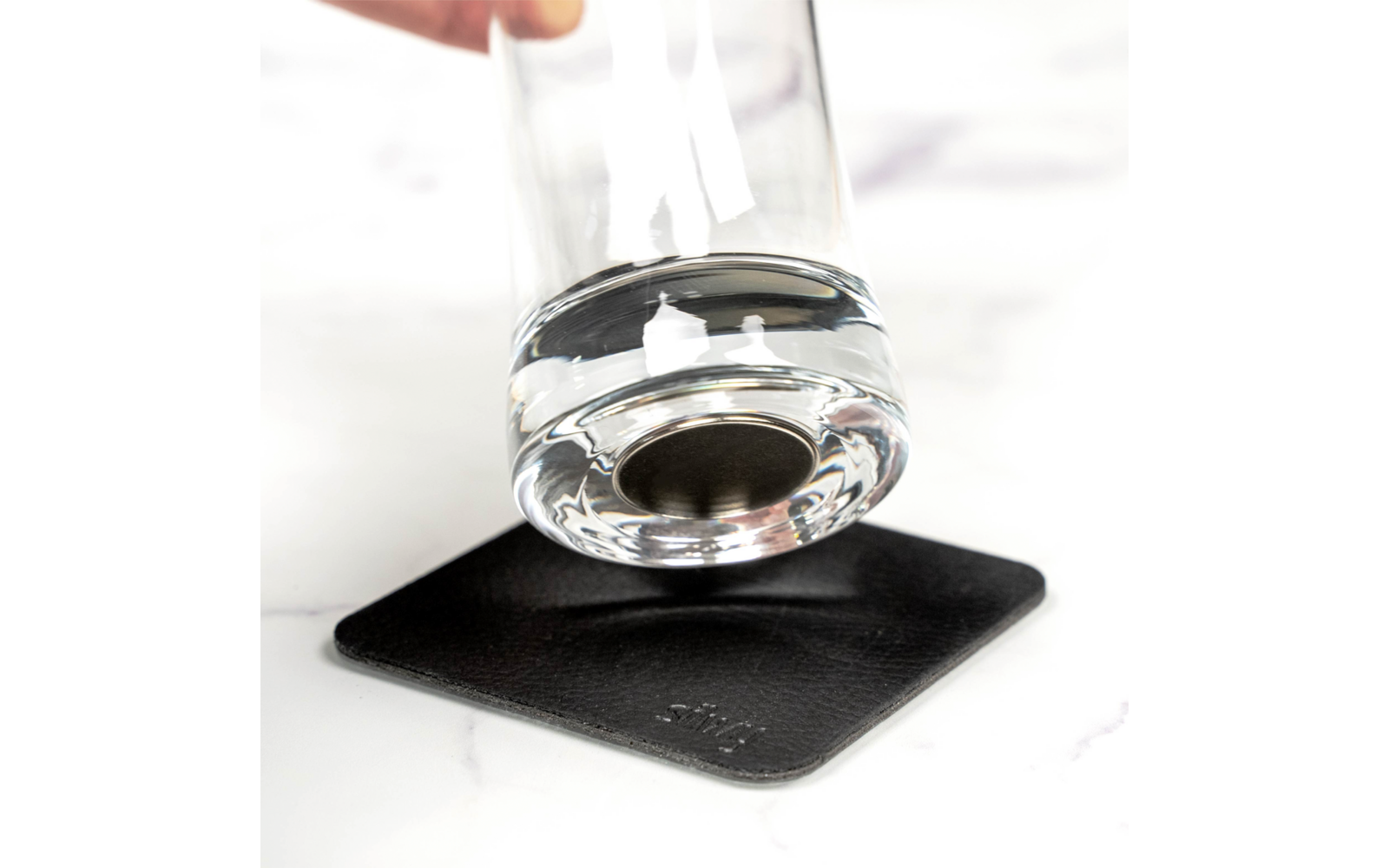 Ein Glas mit Wasser wird über einem schwarzen quadratischen Untersetzer gehalten