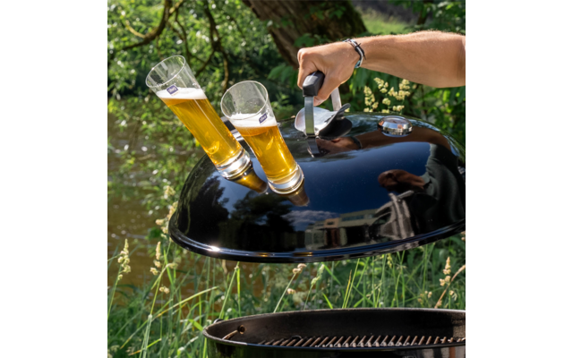 Ein schwarzer Grilldeckel mit zwei darauf befestigten Biergläsern, eine Hand hebt den Deckel an, im Hintergrund grüne Pflanzen