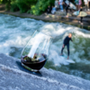 Un verre de vin rouge est posé sur une pierre au bord de la rivière, une personne surfe sur une vague stationnaire en arrière-plan