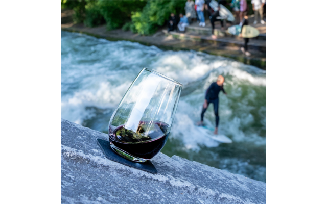 Un verre de vin rouge est posé sur une pierre au bord de la rivière, une personne surfe sur une vague stationnaire en arrière-plan