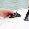 Hand zieht ein schwarzes, dreieckiges Klebeetikett mit weißer, wiederholter Schrift von einer weißen Bootsoberfläche; Wasser im Hintergrund