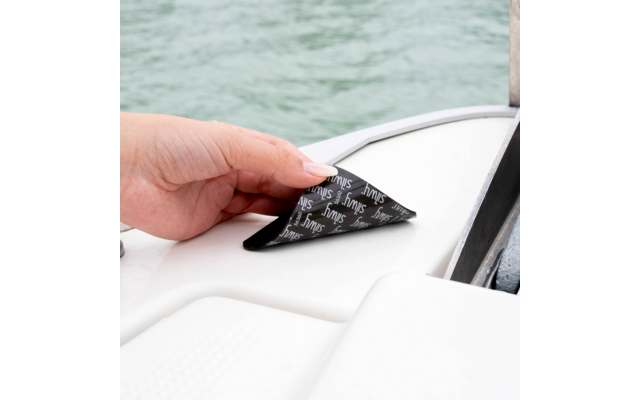 Hand zieht ein schwarzes, dreieckiges Klebeetikett mit weißer, wiederholter Schrift von einer weißen Bootsoberfläche; Wasser im Hintergrund