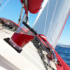 Rode drank in een doorzichtige plastic beker in een ingebouwde bekerhouder op een wit zeilbootdek, rode zeilhoes, touwen en metalen bevestigingen, witte zeilen, blauwe zee en kust op de achtergrond