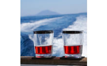Two glasses with red liquid on a railing, in the background a boat wake on the sea and a distant island on the horizon