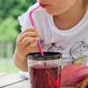Gedeeltelijke weergave van een persoon die via een roze rietje uit een doorzichtig glas met donkerrode drank drinkt; wit T-shirt met print; houten tafel; onscherpe groene achtergrond