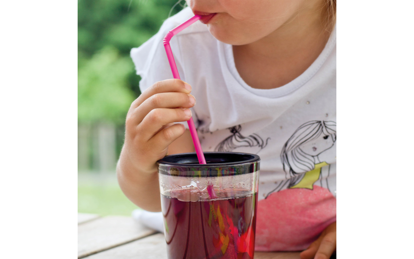 Gedeeltelijke weergave van een persoon die via een roze rietje uit een doorzichtig glas met donkerrode drank drinkt; wit T-shirt met print; houten tafel; onscherpe groene achtergrond