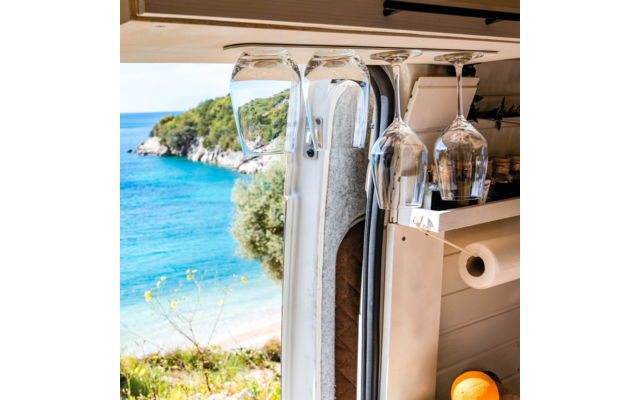 Wine glasses hanging in a camper in front of an open window overlooking the sea and a wooded hill