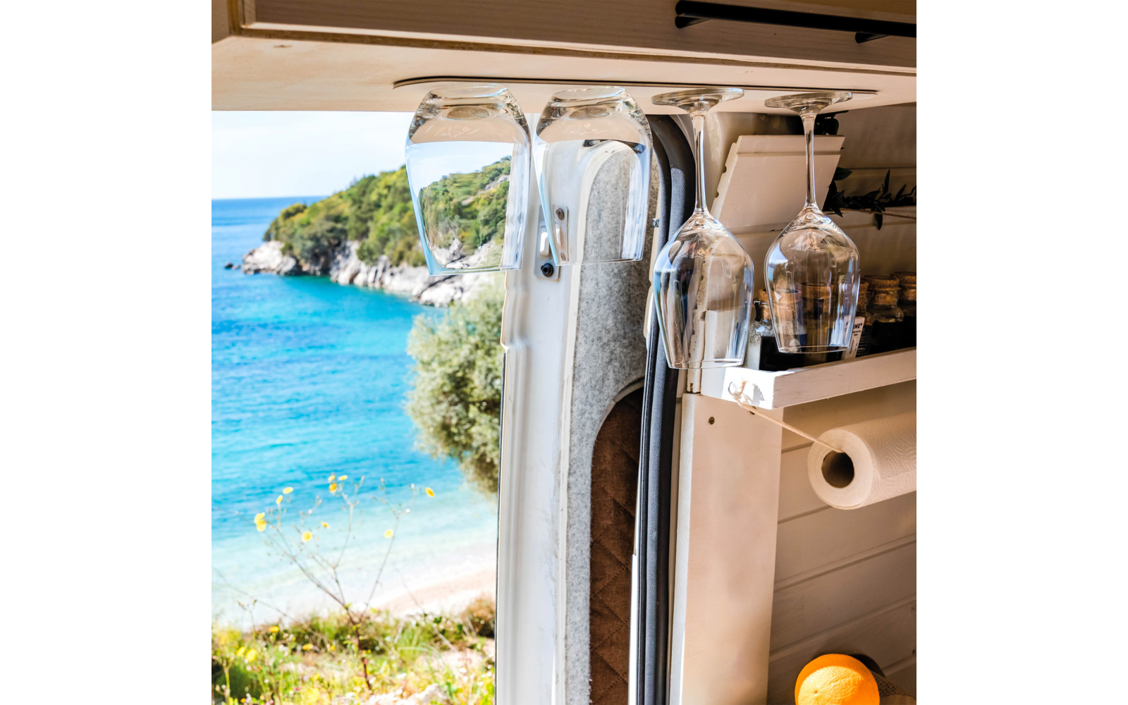 Wine glasses hanging in a camper in front of an open window overlooking the sea and a wooded hill