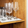 Verschiedene Glasarten, darunter Weingläser und Wassergläser, stehen auf einem weißen Tablett in einem Holzregal