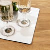 Zwei hohe Gläser mit prickelnder, heller Flüssigkeit auf einer rechteckigen weißen Matte neben einer kleinen weißen Vase mit grünen Blättern auf einem Holztisch
