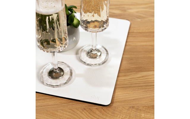 Zwei hohe Gläser mit prickelnder, heller Flüssigkeit auf einer rechteckigen weißen Matte neben einer kleinen weißen Vase mit grünen Blättern auf einem Holztisch