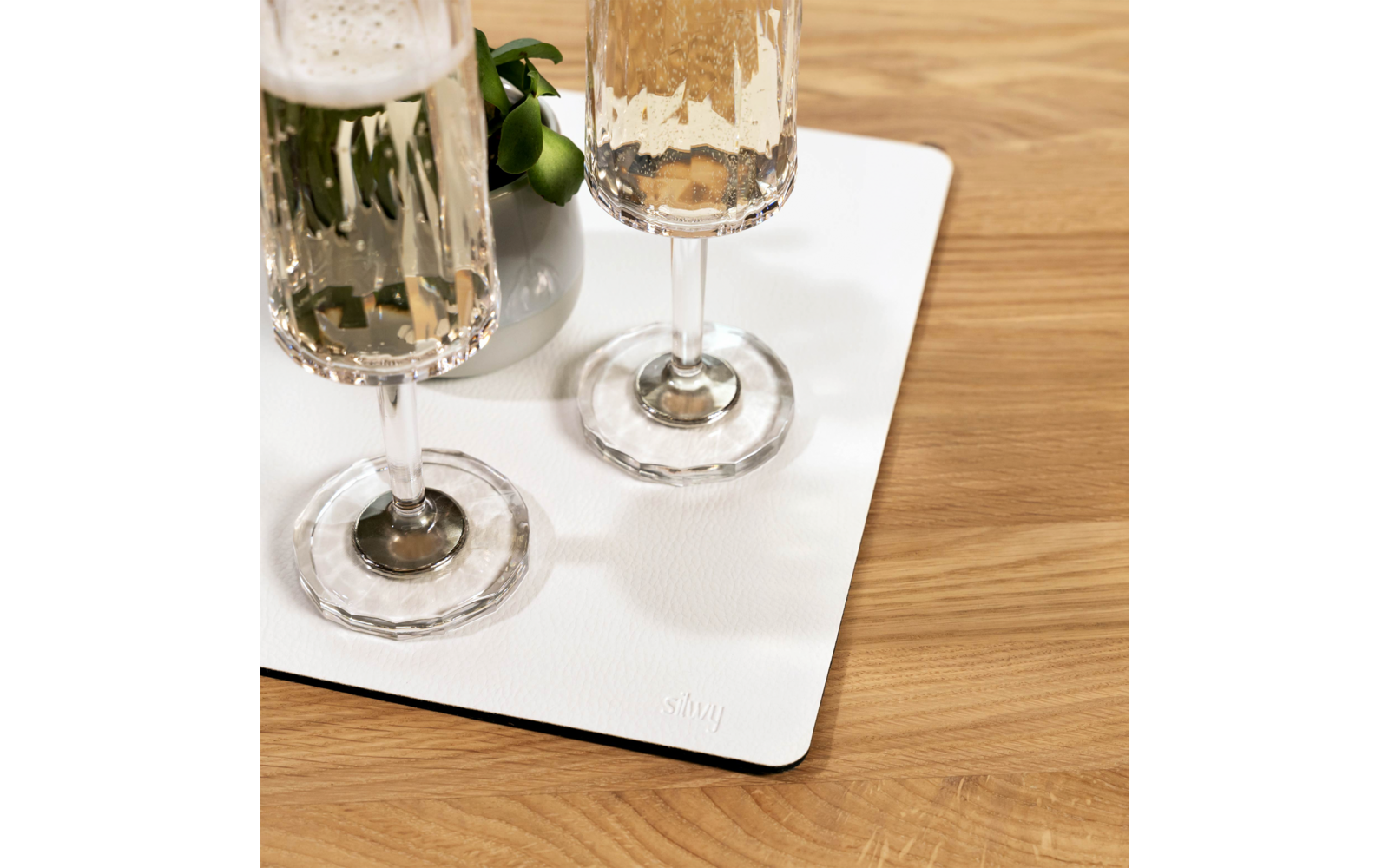 Zwei hohe Gläser mit prickelnder, heller Flüssigkeit auf einer rechteckigen weißen Matte neben einer kleinen weißen Vase mit grünen Blättern auf einem Holztisch