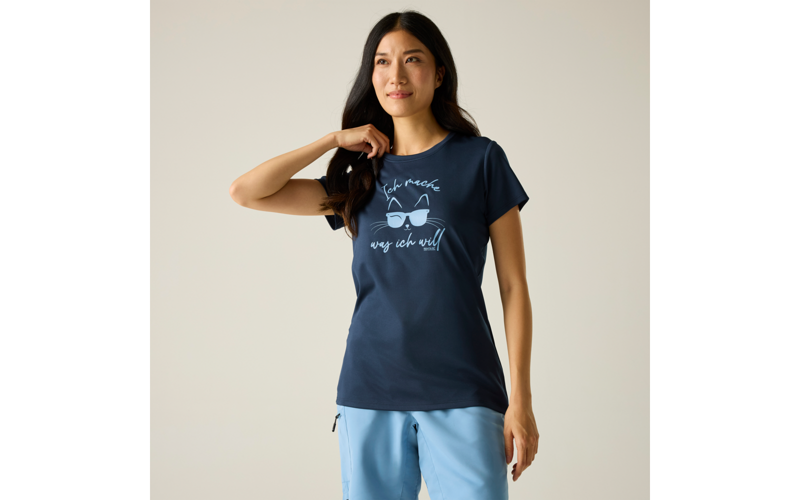 Femme debout devant un fond clair, porte un T‑shirt bleu foncé avec illustration de hibou et texte, pantalon bleu clair, main droite dans les cheveux, regard sur le côté