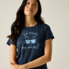 Femme, portrait mi‑corps, cheveux longs foncés, t‑shirt bleu marine avec motif de visage de chat portant des lunettes de soleil et texte « Ich mache was ich will », bras croisés devant le ventre, fond uni clair