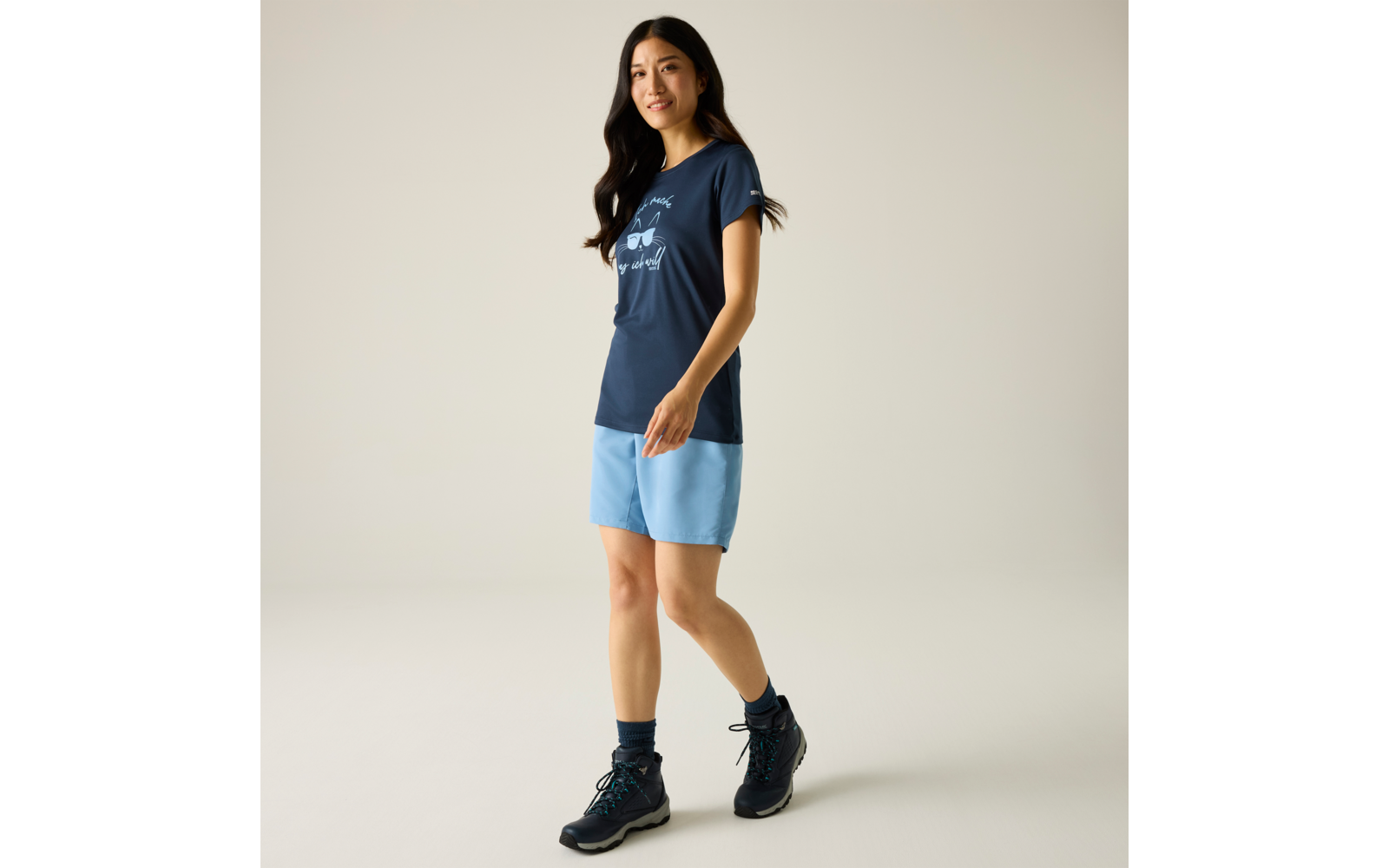 Personne aux longs cheveux foncés, t‑shirt bleu marine avec impression blanche, short bleu clair, chaussures de randonnée foncées et chaussettes foncées, fond uni clair