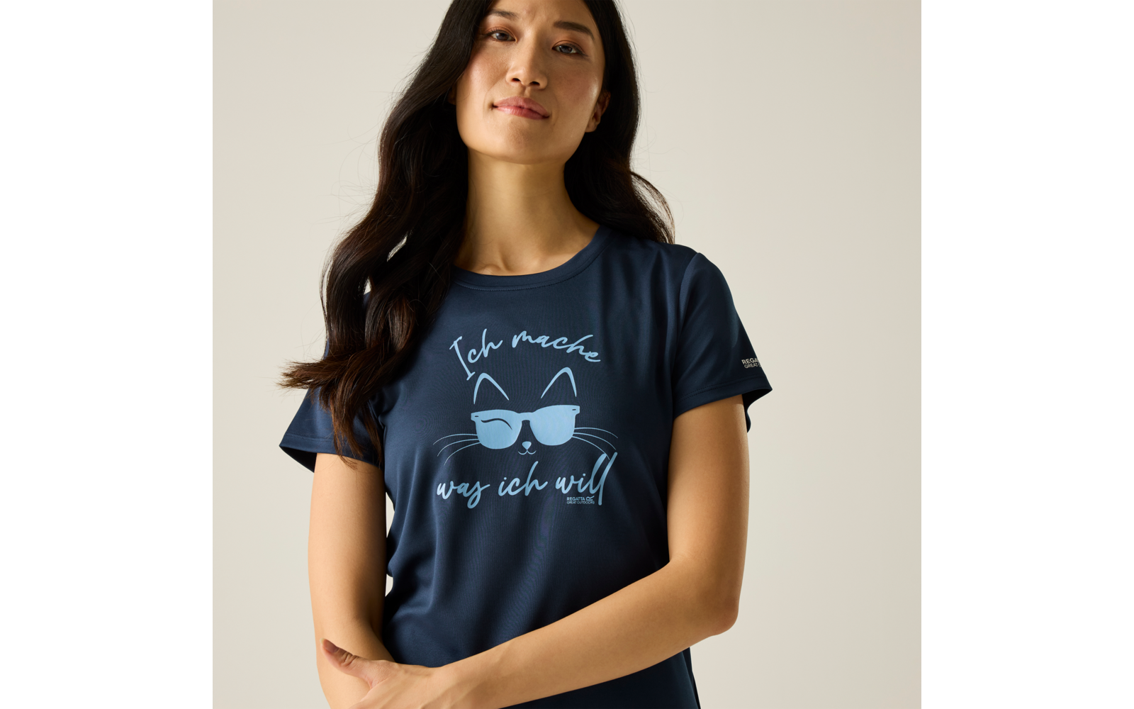 Femme, portrait mi‑corps, cheveux longs foncés, t‑shirt bleu marine avec motif de visage de chat portant des lunettes de soleil et texte « Ich mache was ich will », bras croisés devant le ventre, fond uni clair