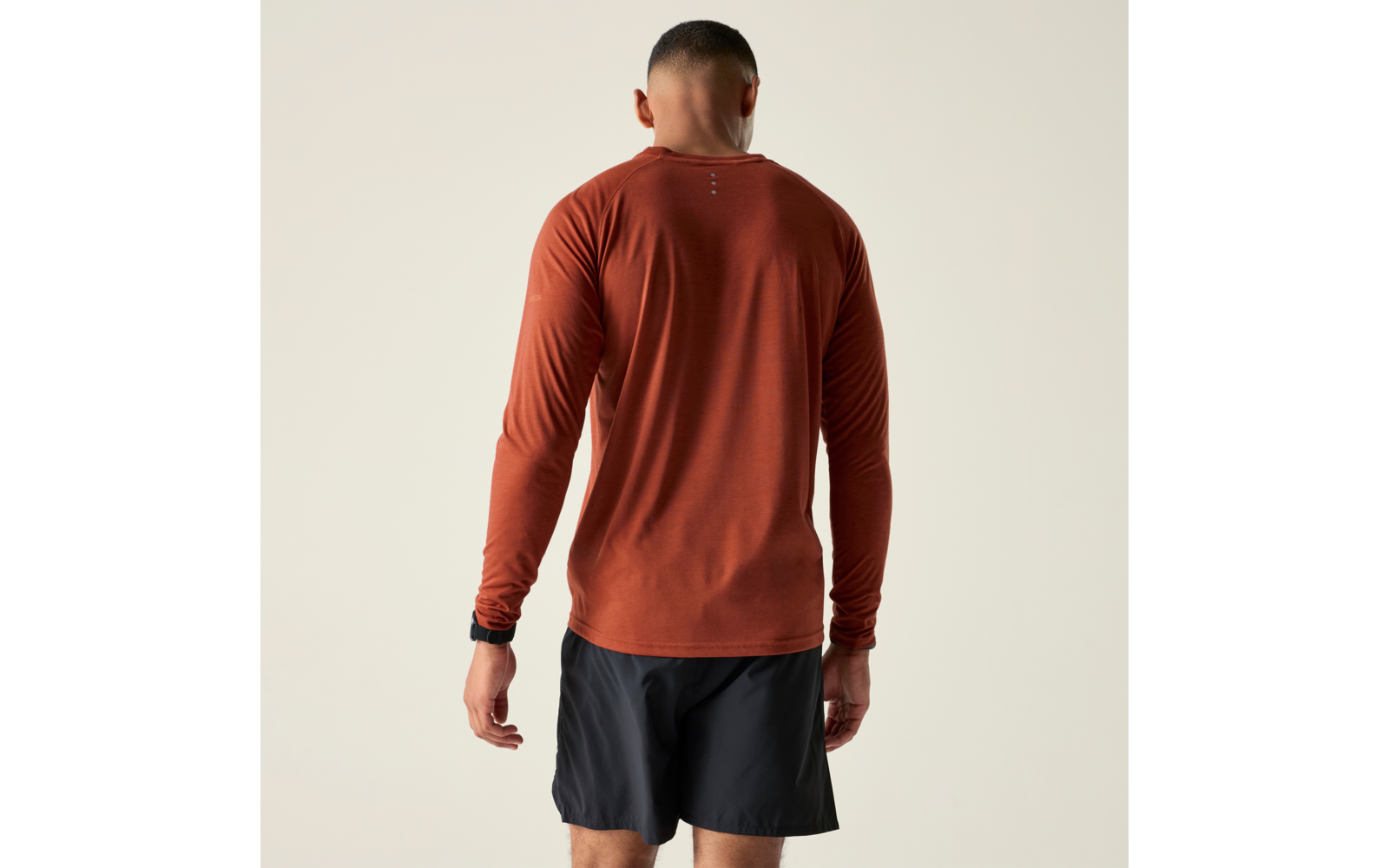 Person von hinten, trägt rostfarbenes Langarmshirt, schwarze Shorts und Armbanduhr, heller Hintergrund