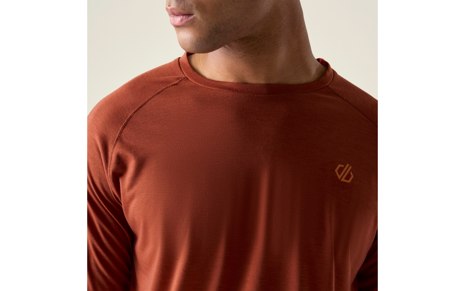 Oberkörper einer Person in rostbraunem Langarmshirt mit kleinem gelb-orangenem Logo auf der linken Brust, unteres Gesicht und Hals sichtbar, Kopf leicht nach rechts gedreht, heller Hintergrund