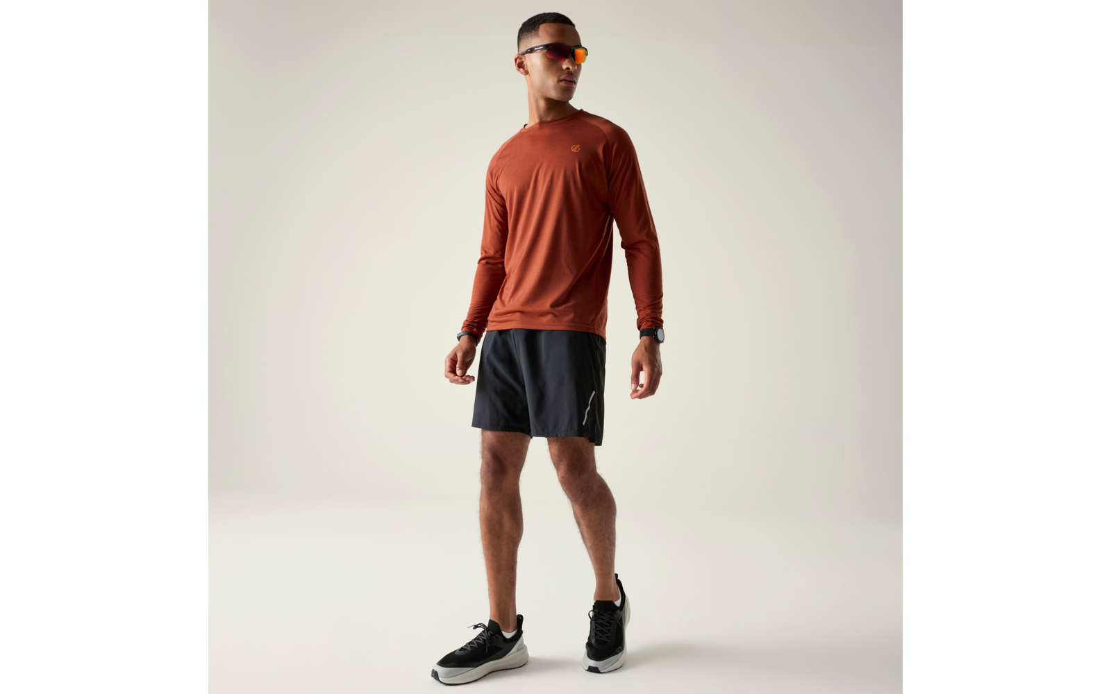 Stehender Mann in rostfarbenem Langarmshirt, schwarzen Sportshorts, schwarzen Sneakern, Sonnenbrille und Armbanduhr vor hellem Hintergrund