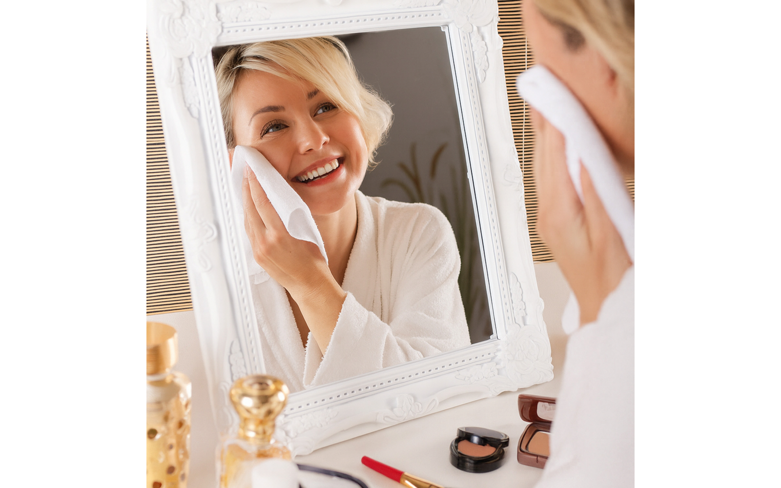 Frau im weißen Bademantel wischt ihr Gesicht mit einem Wattepad vor einem weißen Spiegel; Schminkutensilien und Parfümflakons auf der Ablage