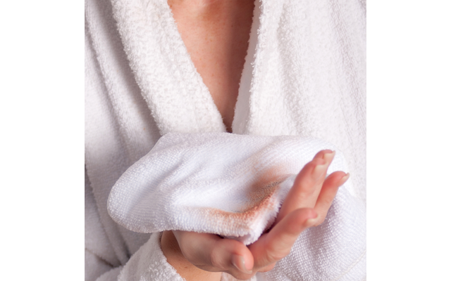 Person in weißem Bademantel hält ein weißes Waschtuch mit bräunlicher Flecken
