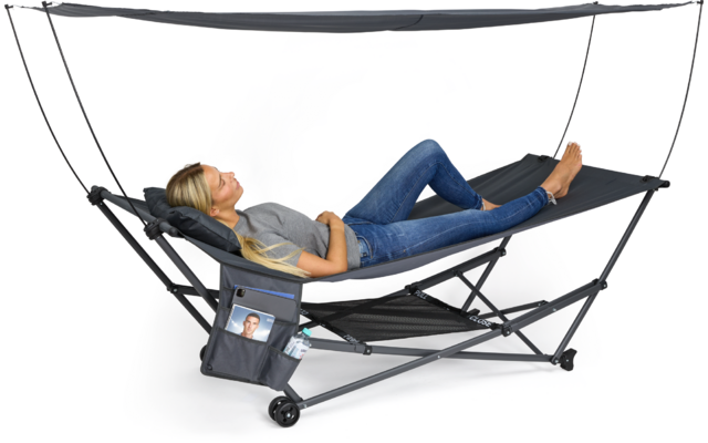 Person mit blondem Haar liegt barfuß auf einer schwarzen, faltbaren Liege mit Metallgestell, Sonnendach und seitlicher Aufbewahrungstasche mit eingestecktem Foto; trägt graues T-Shirt und blaue Jeans