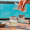 Eine Hand gießt Wasser aus einem Krug in ein Glas auf einem Holztisch neben zwei weiteren Gläsern