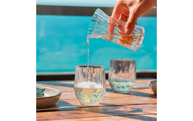 Eine Hand gießt Wasser aus einem Krug in ein Glas auf einem Holztisch neben zwei weiteren Gläsern