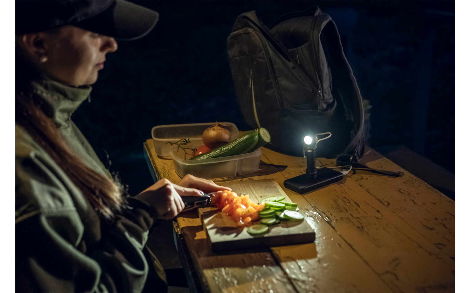Person schneidet Gemüse auf einem Brett, beleuchtet von einer Lampe auf einem Tisch, dunkler Hintergrund