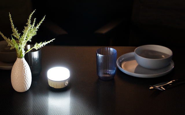 Dunkler Tisch mit leuchtender runder LED-Lampe, weißer Vase mit grünen Zweigen links, blaues Trinkglas daneben, rechts gestapelte graue Schalen und Teller sowie Besteck