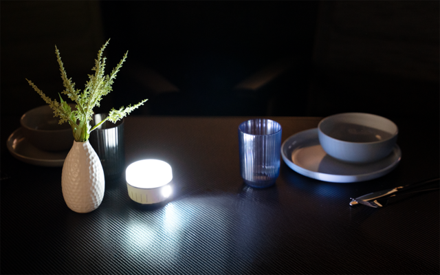 Dunkler Tisch mit weißer Vase mit grünen Zweigen, leuchtendem rundem LED-Licht, blauem Trinkglas, gestapelten Tellern und Schalen, Besteck